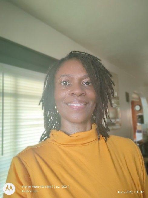 Person wearing a yellow turtleneck sweater standing indoors with windows and furniture in the background.